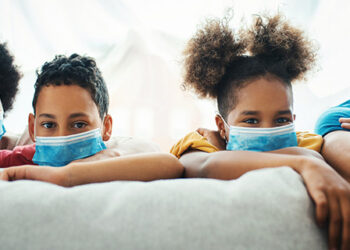 Closeup front view of an african american and hispanic family wearing face masks and staying at home. They are leaning over sofa and looking at the camera.