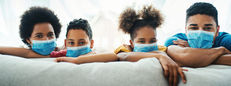 Closeup front view of an african american and hispanic family wearing face masks and staying at home. They are leaning over sofa and looking at the camera.